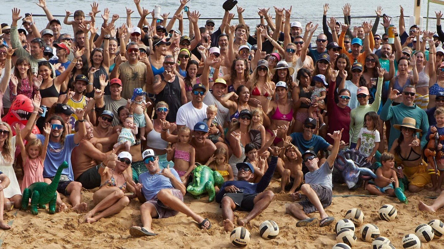 A large group of beach volleyball players, families and children waving together on the sand beside the ocean