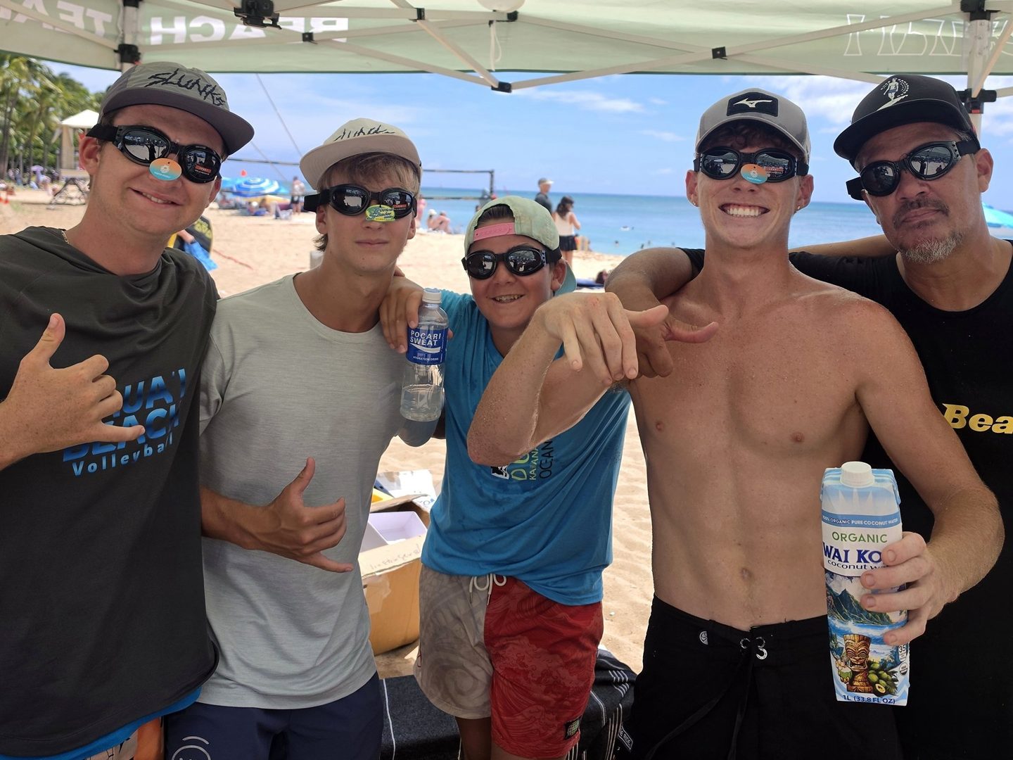 Five community members smiling together beneath a beach tent, with one holding Wai Koko coconut water